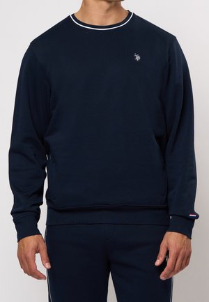 Homme portant un sweat-shirt à manches longues bleu marine et un pantalon assorti bleu marine, avec un petit logo blanc sur la poitrine et le pantalon.