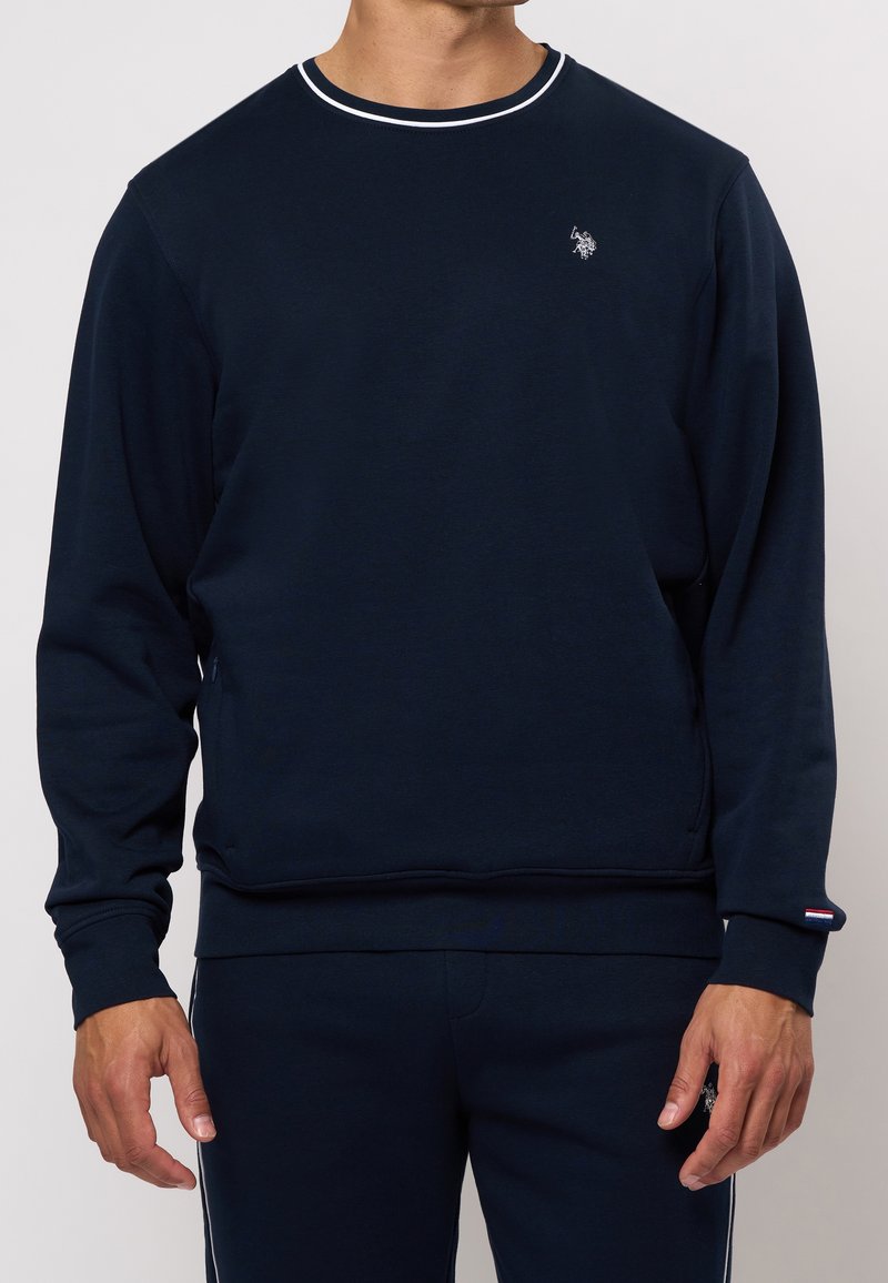 Homme portant un sweat-shirt à manches longues bleu marine et un pantalon assorti bleu marine, avec un petit logo blanc sur la poitrine et le pantalon.
