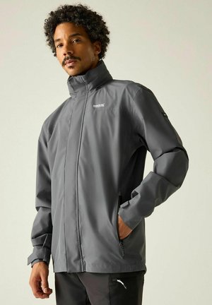 Man met krullend haar die een grijze Regatta waterdichte jas draagt met hoge kraag, ritssluitingen in de zakken en een zwarte broek tegen een lichte achtergrond.
