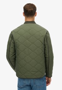 Olijfgroene doorgestikte jas met een geribde kraag en contrasterende zijstrepen, voorzien van een horizontaal golfpatroon voor textuur.