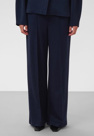 Personne portant un pantalon large bleu marine foncé et une veste assortie, debout sur un sol gris clair, les mains détendues le long du corps.