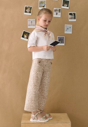 Ragazza giovane con pantaloni a fantasia e maglietta bianca in piedi su una cassa, che tiene una foto con diverse foto esposte sulla parete dietro di lei.