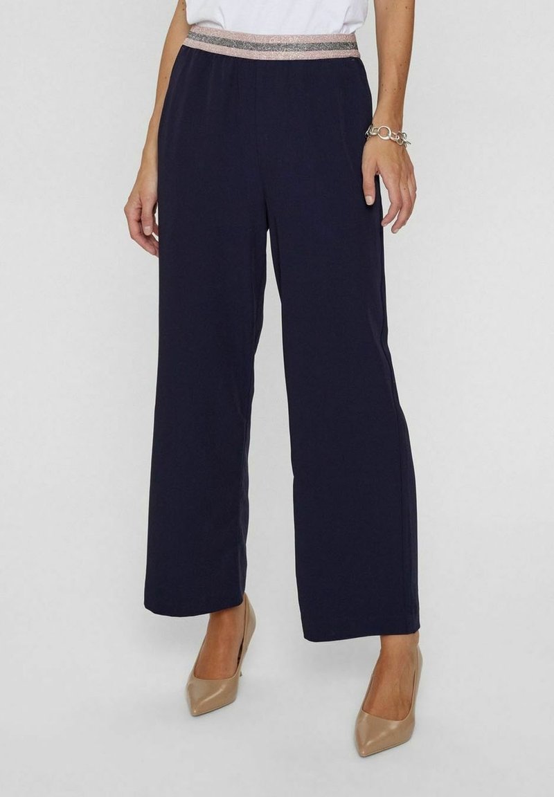 Pantalon large bleu marine pour femme avec taille élastique métallique, associé à des talons pointus beiges et un top blanc.
