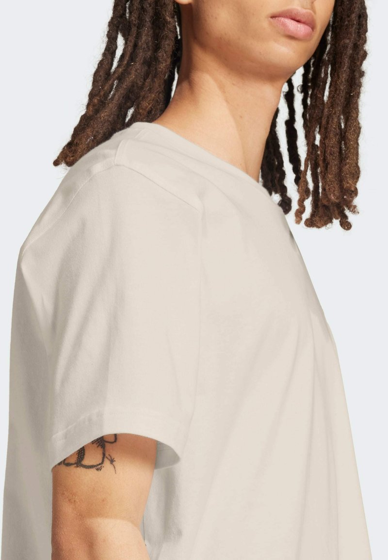 Persona con rastas largas que lleva una camisa suelta de manga corta color blanco roto, mostrando un tatuaje en la parte superior del brazo sobre un fondo liso.