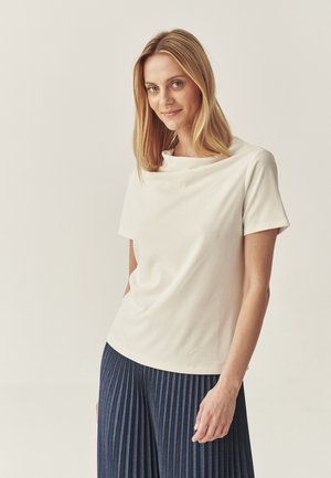Cream-colored short-sleeve top with a draped neckline, paired with dark pleated pants. Smooth fabric with a soft texture.