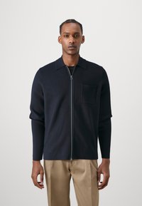 Navy zip-up knit cardigan with a collar and a chest pocket, featuring long sleeves and a smooth texture, paired with beige trousers.