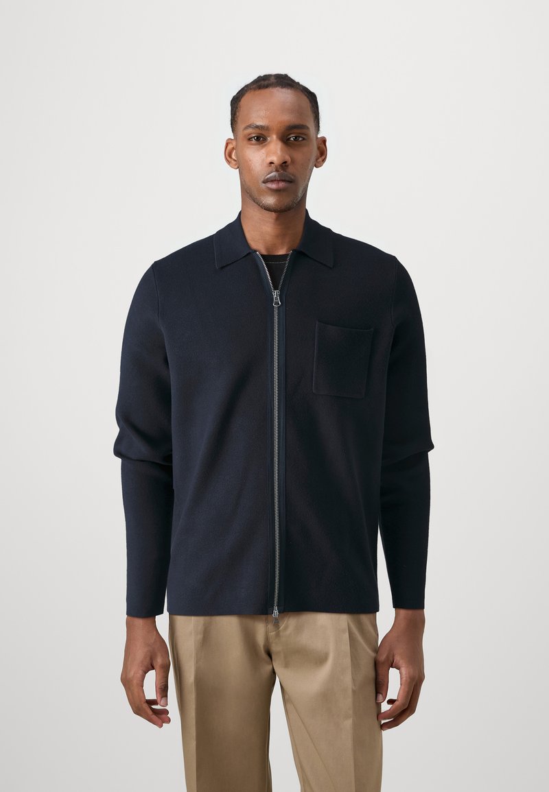 Navy zip-up knit cardigan with a collar and a chest pocket, featuring long sleeves and a smooth texture, paired with beige trousers.