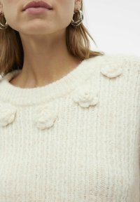 Kreemist kudumistest sweater, millel on ribakujuline tekstuur, lillede applikaadid kaeluses ja ümmargune kaelus.