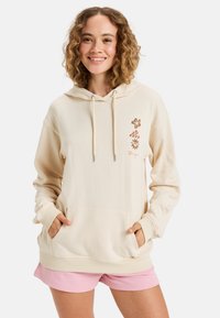 Beige hoodie met een capuchon, verstelbare koordjes, een voorke pocket en bloemenpatronen in bruin aan de linkerkant; gecombineerd met roze shorts.