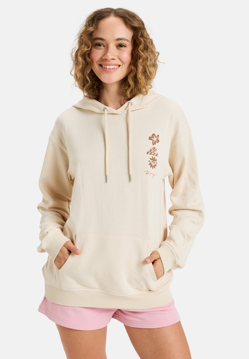 Beige hoodie met een capuchon, verstelbare koordjes, een voorke pocket en bloemenpatronen in bruin aan de linkerkant; gecombineerd met roze shorts.