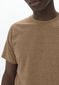 Brown cotton t-shirt with a crew neck, featuring a textured surface and short sleeves, partially shown on a person.