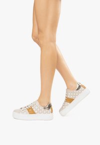 Sneaker color sabbia chiaro con soletta bianca, caratterizzate da una tomaia a motivo, dettagli dorati e un tacco metallico. I lacci sono bianchi e piatti.