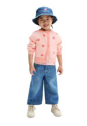 Jeune enfant portant un cardigan rose à motifs fraises, un jean bleu à jambes larges, des baskets blanches et un chapeau bob bleu, souriant et debout.