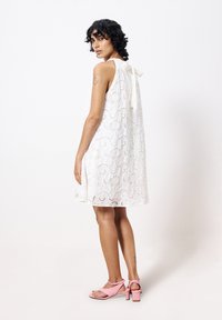Weißes Spitzenkleid mit ausgestelltem Schnitt, ärmellosem Design und Schlaufen-Detail auf der Rückseite. Kombiniert mit rosa Absatzsandalen.