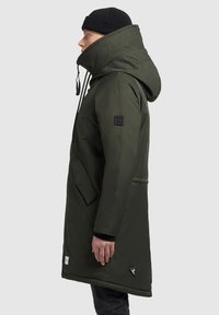 Man in zwarte beanie met een donkergroene gewatteerde winterparka met hoge kraag, staande in profiel tegen een effen lichte achtergrond.