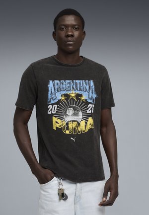 Młody mężczyzna noszący wyblakły czarny T-shirt Argentina Puma z grafiką piłki nożnej i gwiazd, jasne dżinsy oraz klucze przypięte do pętli paska.