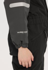 Veste extérieure noire avec manches grises, présentant le logo "W-PRO 10000" et des poignets élastiques. Tissu doux avec des détails de poches latérales zippées.