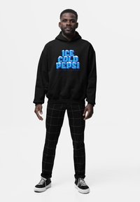 Uomo in piedi che indossa un felpa nera con la scritta "Ice Cold Pepsi" e pantaloni a scacchi neri, su sfondo bianco.