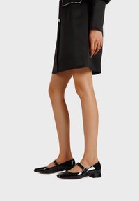 Scarpe Mary Jane in pelle verniciata nera con tacco basso, dotate di una cinturino decorativo e dettaglio fibbia. Finitura liscia e lucida.