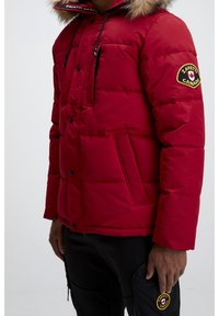 Veste matelassée rouge avec capuche bordée de fourrure, présentant un patch logo, une fermeture éclair à l'avant et deux poches latérales. Texture de tissu lisse et matelassée.