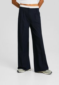 Marineblauwe gestreepte wijde broek met een elastische witte tailleband, met een ontspannen pasvorm en subtiele geplooide details, gecombineerd met lichtgekleurde sneakers.