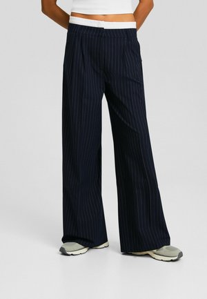 Pantalons larges à rayures marines avec une ceinture élastique blanche, offrant une coupe décontractée et des plis subtils, accompagnés de baskets de couleur claire.