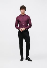 Bordowa koszula na guziki z długim rękawem i kołnierzykiem, zestawiona z czarnymi dopasowanymi spodniami i czarnymi sznurowanymi butami, stojąca na białym tle.