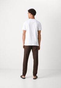 Camiseta de algodón blanca con mangas cortas, combinada con pantalones marrones ajustados y chanclas negras, vista desde la parte posterior. Diseño sencillo, sin patrones.