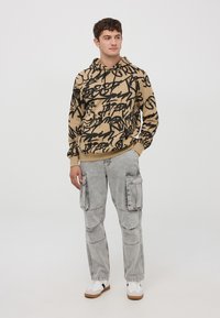 Felpa beige con stampa in stile graffiti nera, maniche elastiche e tasca frontale. Abbinata a pantaloni cargo grigio chiaro con molteplici tasche.
