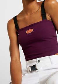 Top cropped viola con spalline nere regolabili e un badge logo arancione al centro. Tessuto di cotone morbido, design aderente.