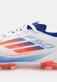 Botas de fútbol blancas con acentos en azul y naranja, con una parte superior texturizada, diseño a rayas, cordones blancos y tachones naranjas en la suela.