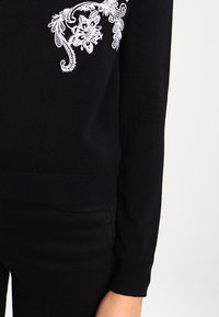 Pull noir en maille avec broderie florale blanche sur l'épaule gauche, poignets côtelés et taille ajustée.