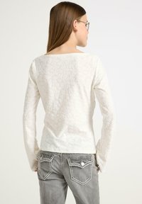 Weißes, strukturiertes Langarmoberteil mit quadratischem Ausschnitt, kombiniert mit grauen Jeans mit dekorativen Gesäßtaschen und kontrastierenden Nähten.