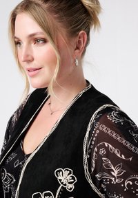 Zwarte suède vest met witte bloemengestikte, gelaagd over een doorschijnend zwart gebloemd blouse. Zilveren oorbellen en ketting voegen accenten toe.