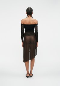 Melna off-the-shoulder blūze ar mežģīņu piedurknēm, apvienota ar caurspīdīgu brūnu apvilkuma svārku ar asimetrisku apakšmalu.