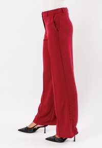 Pantaloni a gamba larga rossi realizzati in tessuto liscio, con tasche frontali, una vita strutturata e un design delle cuciture pulito. Indossati con tacchi neri.