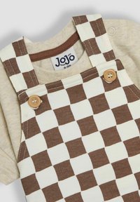 Pantalones de tirantes a cuadros marrón y crema con botones de madera, sobre una blusa de color beige claro. La tela parece suave con un diseño casual.