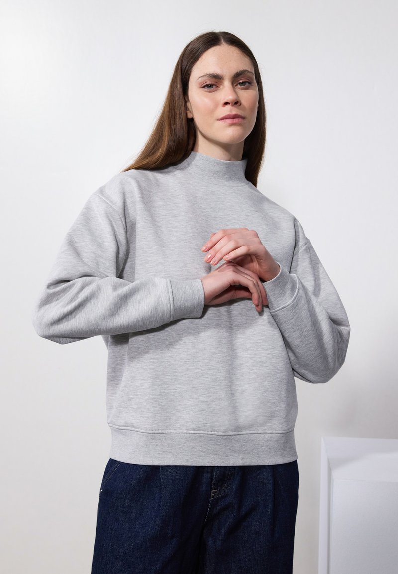 Sudadera gris clara con cuello alto y puños acanalados. Diseño ajustado, textura suave y una silueta relajada combinada con pantalones de mezclilla oscuros.