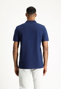 Polo blu navy con colletto classico e maniche corte, realizzato in tessuto liscio, mostrato da dietro con pantaloni bianchi.