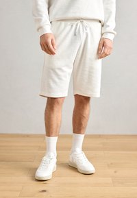 Hvite teksturerte shorts med elastisk snøring i livet, kombinert med hvite ankelstrømper og hvite joggesko, på et tregulv.