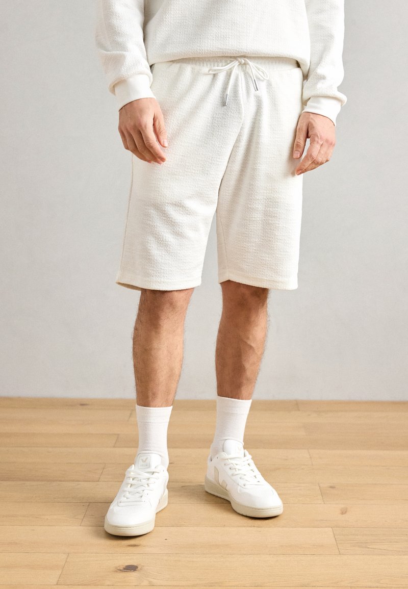Hvite teksturerte shorts med elastisk snøring i livet, kombinert med hvite ankelstrømper og hvite joggesko, på et tregulv.