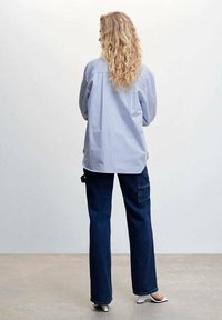 Blauw en wit gestreepte shirt met lange mouwen, losse pasvorm; gecombineerd met donkerblauwe flared jeans en witte heeled mules. Model staat met de rug naar de camera.