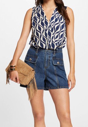Vrouw draagt een blauw-witte mouwloze blouse met patroon, hooggetailleerde denim shorts met zakken, houdt een beige suède clutch met franjes en gouden armbanden vast.