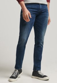 Jeans de mezclilla azul oscuro con corte ajustado, que presentan un ligero desgaste y un diseño clásico de cinco bolsillos, combinados con zapatillas altas en blanco y negro.