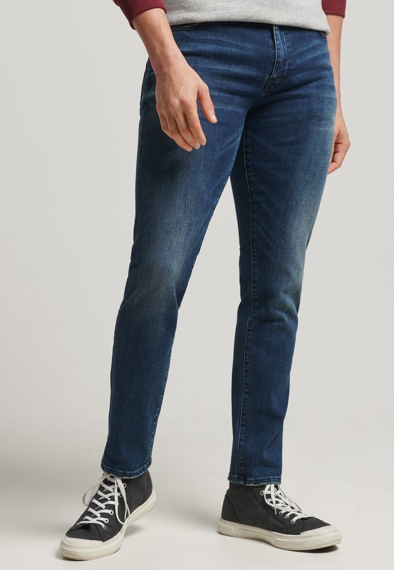 Jeans de mezclilla azul oscuro con corte ajustado, que presentan un ligero desgaste y un diseño clásico de cinco bolsillos, combinados con zapatillas altas en blanco y negro.