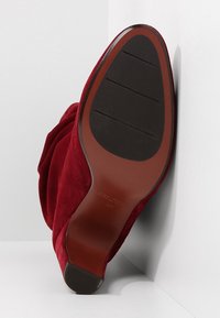 Bota de ante roja con punta redondeada y diseño arrugado. La suela es marrón con líneas de tracción negras. Etiqueta de tamaño visible en el interior.