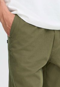 Hand resting in pocket of olive green pants worn with a white shirt, focusing on midsection and fabric texture.