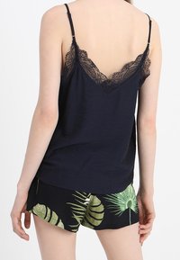 Navyfarbene Spaghetti-Top mit Spitzenbesatz auf der Rückseite, kombiniert mit schwarzen Shorts mit grünem Blattmuster. Glattes Material, verstellbare Träger.