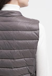 Gilet grigio imbottito con design a righe orizzontali, leggera lucentezza e collo alto, indossato sopra una maglietta bianca a maniche lunghe, mostrando una texture liscia.