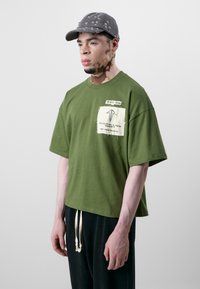 Oliven grønn oversized t-skjorte med lommepatch, hvite etikettgrafikker og kortermet, kombinert med svarte bukser med snøring.